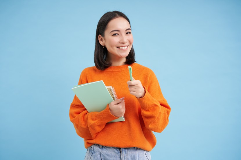 Education and students. Happy asian woman, holding notebooks and laughing, smiling at camera, enjoys going to University or College, blue background.