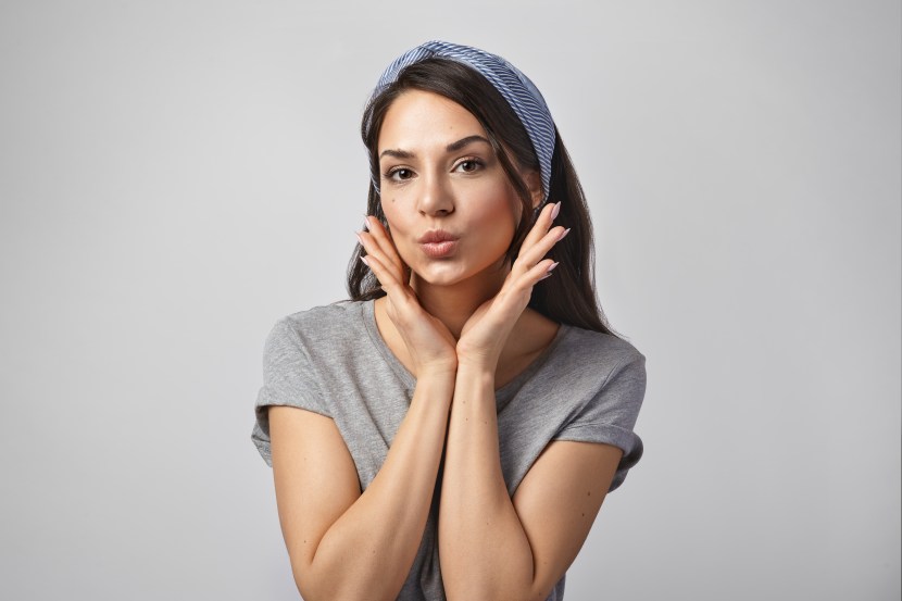 Portrait of coquettish young lady with long dark hair and tanned perfect skin wearing headband looking at camera with flirty expression, pouting lips as if giving kiss, holding hands at her face