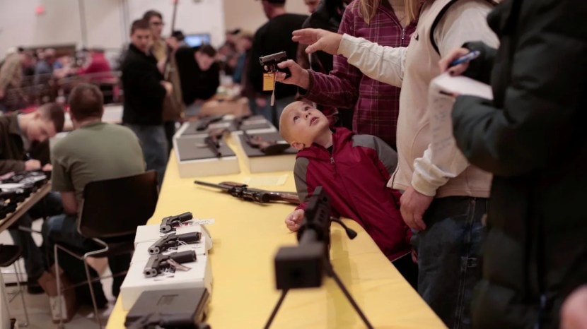 A boy looks on as a customer inspects a pistol at the Saratoga Arms Fair at the City Center in Saratoga Springs, N.Y., Jan. 12, 2013. The New York Times