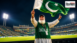 A smiling elderly Pakistani man with a long white beard and black cap, wearing a traditional white outfit and a green vest with Pakistan flag logos, waves a large Pakistan flag enthusiastically under bright floodlights in a packed cricket stadium at night; एका दाढीवाल्या वृद्ध पाकिस्तानी व्यक्तीने क्रिकेट स्टेडियमच्या फ्लडलाईट्समध्ये पाकिस्तानचा मोठा ध्वज उत्साहाने फडकवतानाचा फोटो. त्याने हिरवा सदरा घातला आहे ज्यावर पाकिस्तानचे ध्वज आहेत.