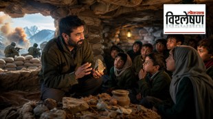 A teacher holds a stone while teaching a group of children inside a rocky cave used as a makeshift classroom, with soldiers and sandbags visible outside against a mountainous backdrop.